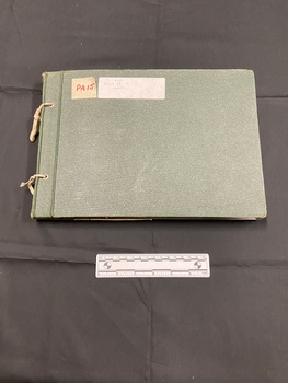 Green cloth bound photo album with two holes punched down the left side and tied with worn elastic. Large white sticker label towards the top that has WWII written on it in black ink.