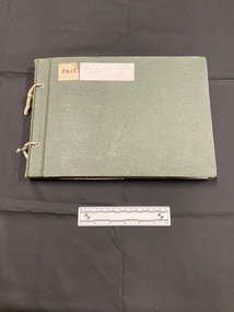 Green cloth bound photo album with two holes punched down the left side and tied with worn elastic. Large white sticker label towards the top that has WWII written on it in black ink.