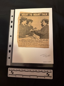Newspaper clipping with a photograph of a woman holding a heart, talking to two young women.