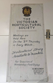 A white tin singe with a red and yellow insignia with the letters RHS at the top. The text on the singe reads Established 1859. RSV. The Victorian Horticultural Society. Meetings are held here on the 3rd Thursday in every month. Horticultural Library Available to Members. For Enquires of Membership & Floral Displays D. Moloney. 276 Canterbury Rd, Heathmont 3135. 7294713 