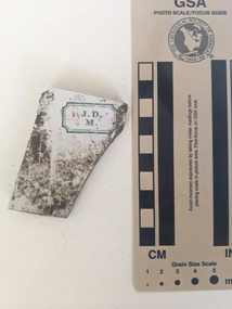 Ceramic shard with white glazed finish and a blue design with the letters J D M