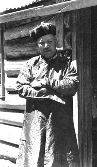 an older man in Mandarin robes stands in front of a hut