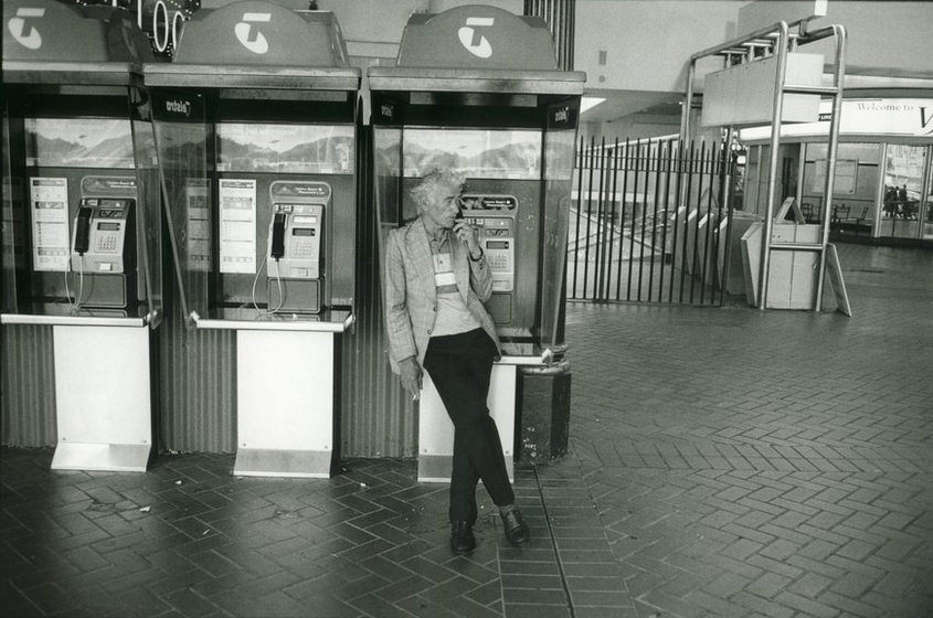 a man leans against a telephone booth whilst smoking a cigarette