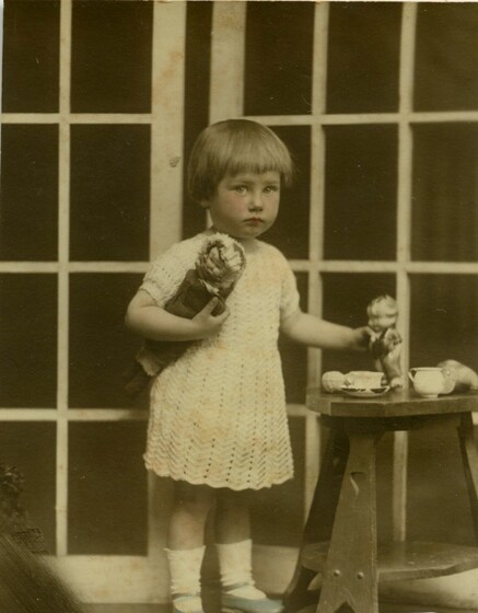 a child in white lace dress stands next to table and tea set, holding two dolls