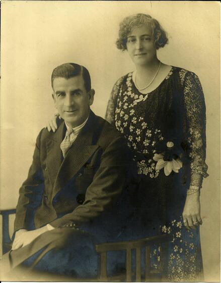 a seated man with woman standing behind, both dressed formally