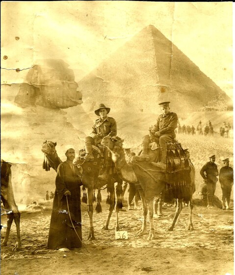 two soldiers on camels are led across the sand with a pyramid in background