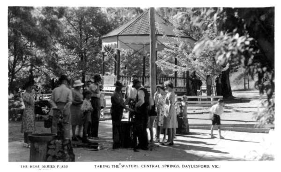 a group of people in the park around a water pump Taking the waters, Central Springs, Daylesford