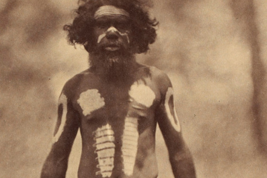 an early photograph, sepia with age, of a man standing on a grassed area wearing ceremonial woven string waist-band and ornamental body paint