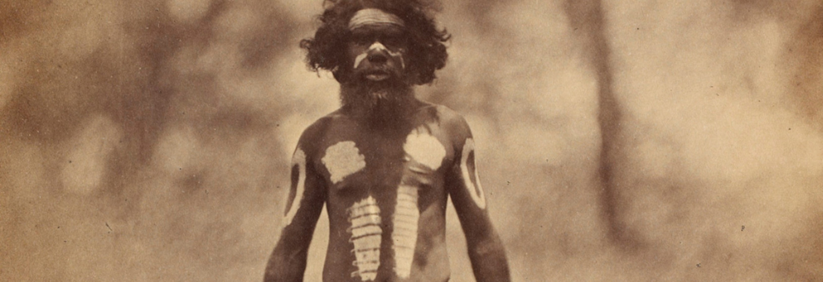 an early photograph, sepia with age, of a man standing on a grassed area wearing ceremonial woven string waist-band and ornamental body paint