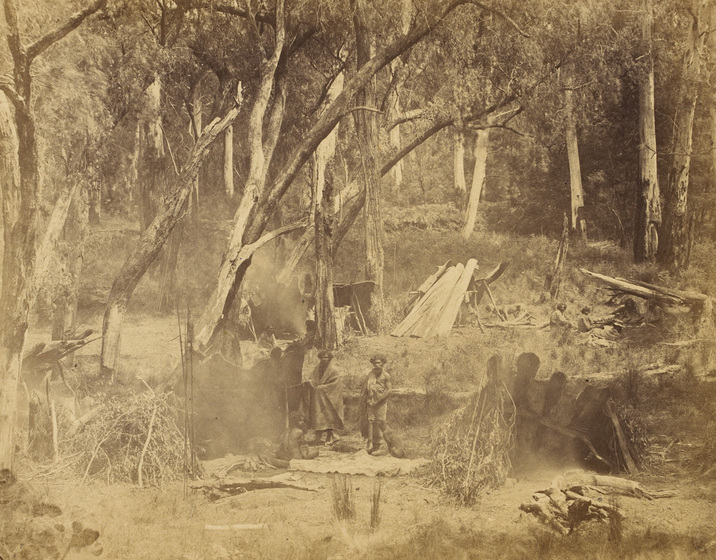 An early photograph, sepia with age, featuring two groups of aboriginal men and women sitting and standing around two fires and several lean-to huts in a treed bush area.