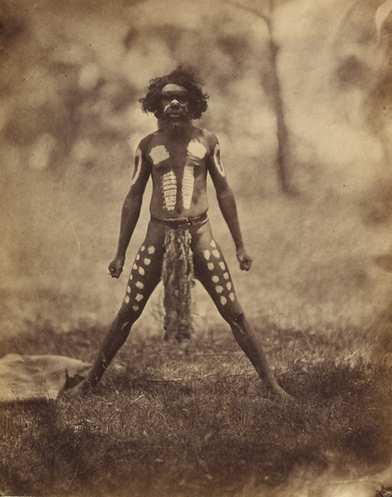 an early photograph, sepia with age, of a man standing on a grassed area wearing ceremonial woven string waist-band and ornamental body paint.