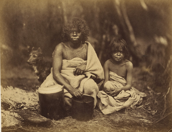 An early photograph, sepia with age, of a woman and child wrapped in blankets sitting beside a fire and holding two billy cans with a dog in the background.