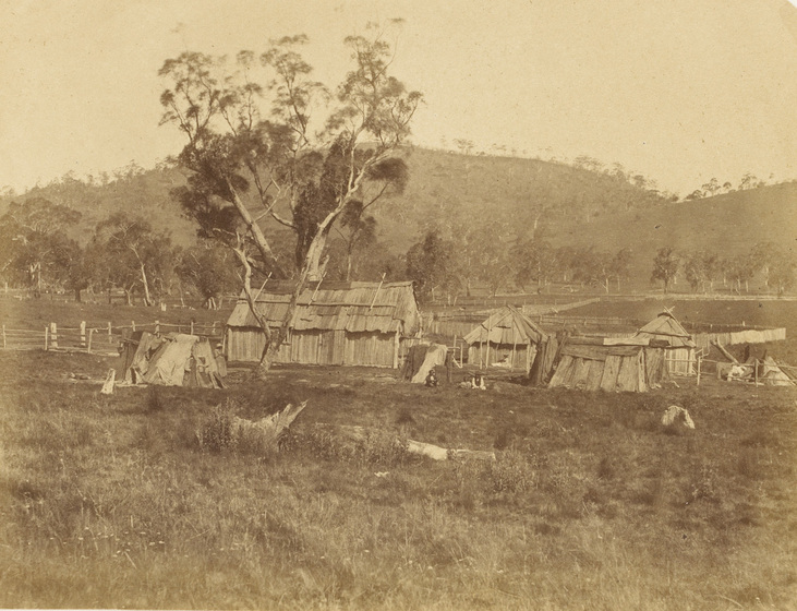 An early photograph, sepia with age, featuring a cluster of six old timber buildings in a rural field.