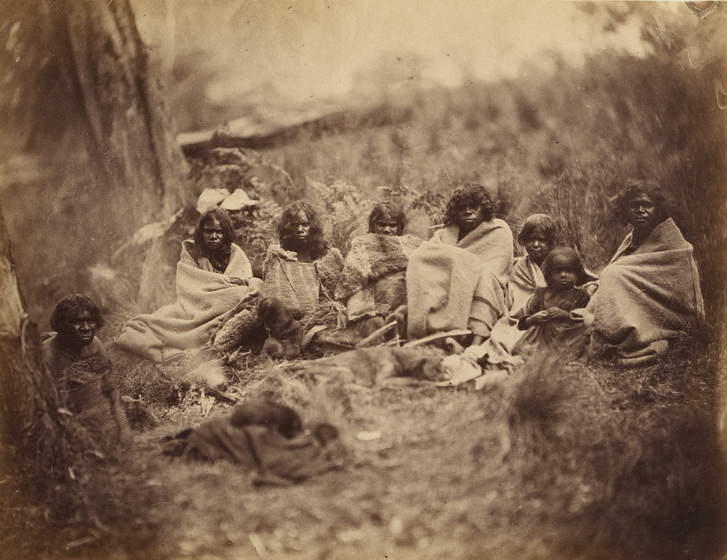 An early photograph, sepia with age, of 9 women and children sitting in a semi-circle on a grassed area with trees.