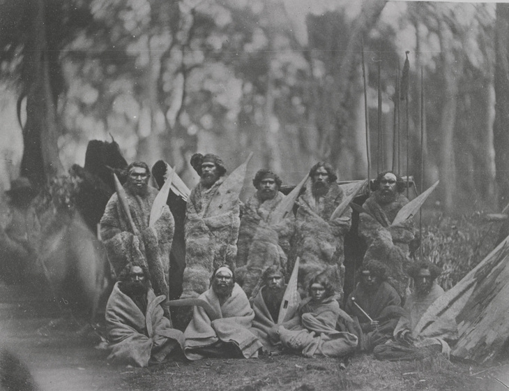 An early photograph of 11 men wrapped in kangaroo skin cloaks and blankets, holding shields and boomerangs with a cluster of spears standing behind. 