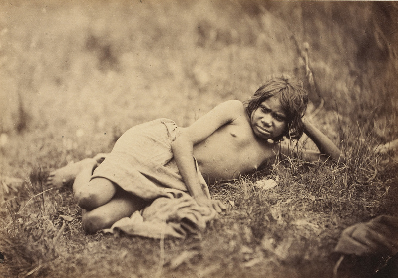 An early photograph, sepia with age, of a young girl reclining in the grass wearing a cloth skirt. 
