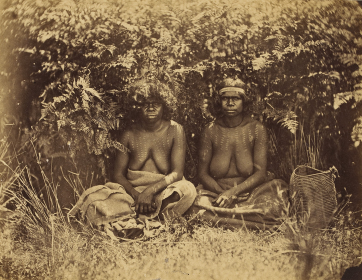 An early photograph, sepia with age, of two women sitting in the shade of bushes with dilly bags beside them on the grass