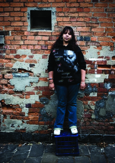 a casually dressed young woman stands against a brick wall looking toward the camera