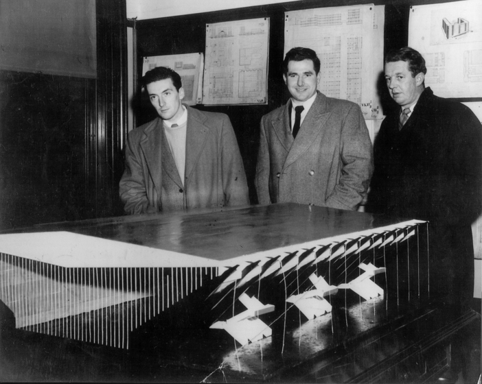 Men standing around a table with a model building on top