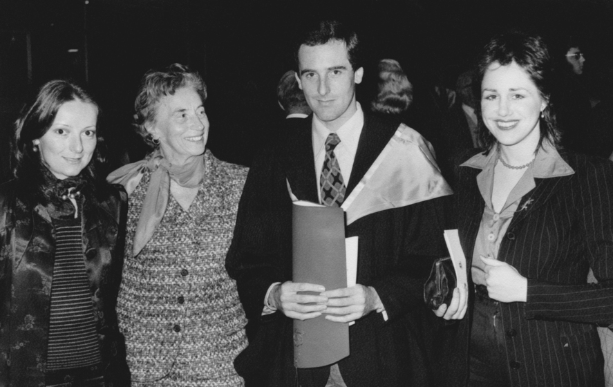 Four people standing next to each other, one wearing graduation regalia