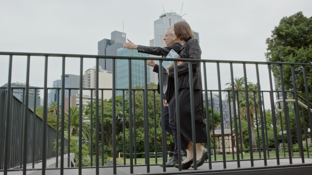 A man and woman walk across a path with metal fencing either side. Behind can be seen trees and tall buildings.