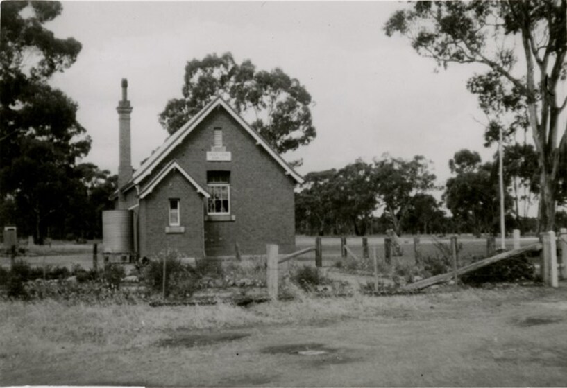 Small double-fronted brick building, with chimney and water tank, in a rural setting with a modest garden and front picket fence.
