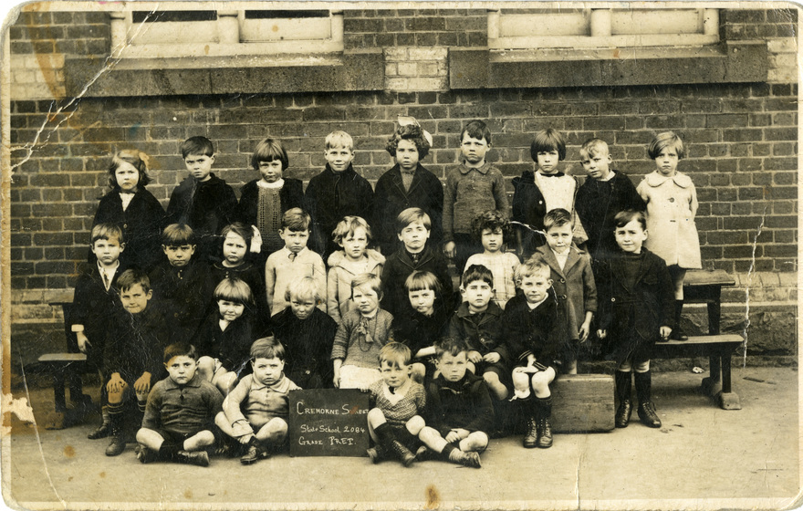 An early black and white photograph of school children, lined-up in four rows, some standing and some seated with a blackboard citing the school name at front centre.