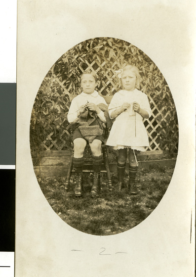 Early photograph of two children, a girl standing and a boy sitting, as they knit items.