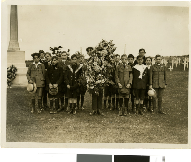 Seventeen young schoolboys, dressed in various  military uniforms stand in the Villers-Bretonneux war cemetery beside the Cross of Sacrifice and carry a large memorial wreath.