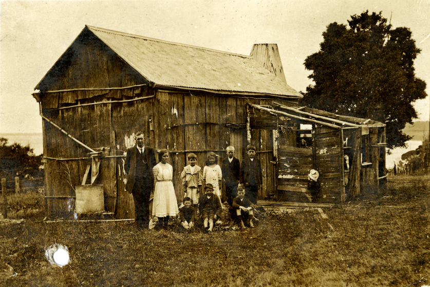 An early photograph of a male teacher and seven pupils of various ages standing outside of a dilapidated single room building representing their school.