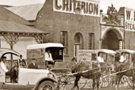 a car and several horse driven carriages in front of brick factory