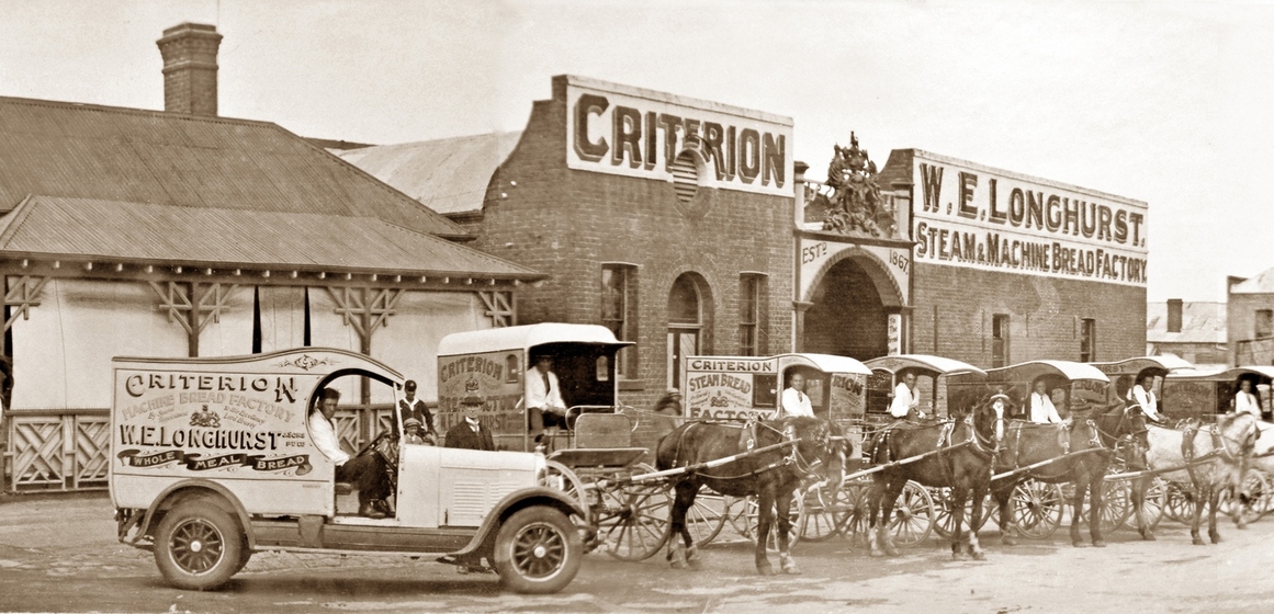Car and several horse driven carriages in front of brick factory building