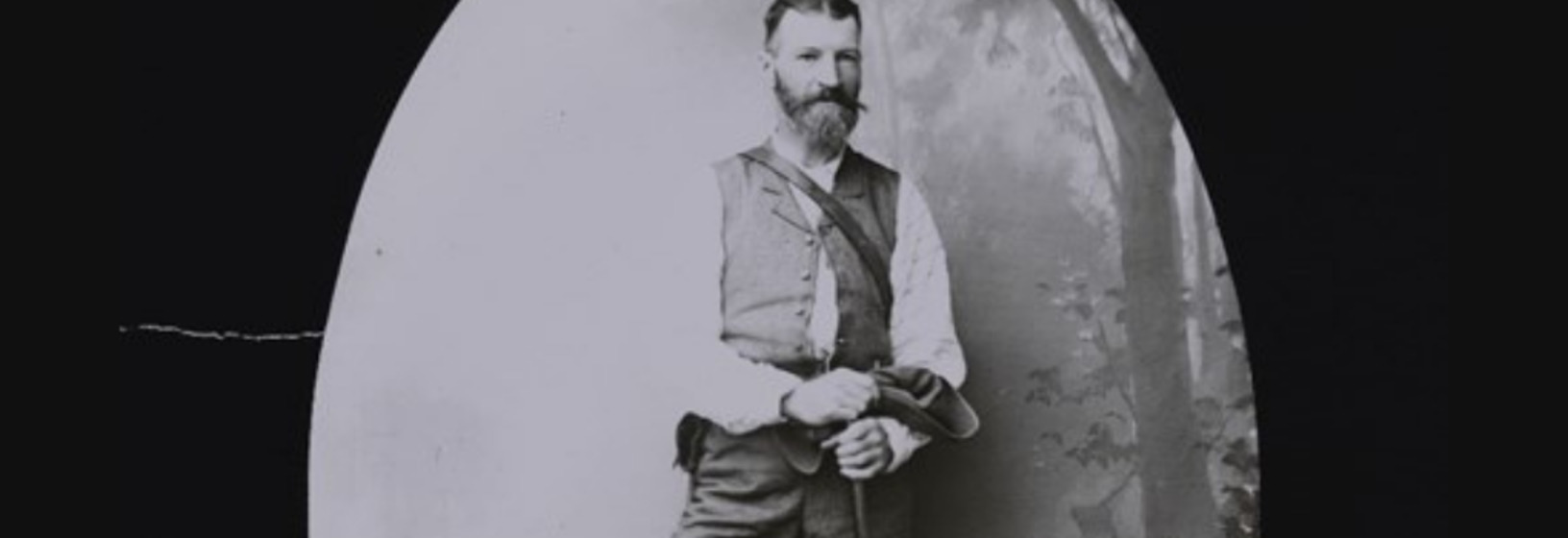 a smartly-dressed man with a beard standing in a forest-scene