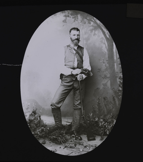 a smartly-dressed man with a beard standing in a forest-scene holding his hat and rifle. 