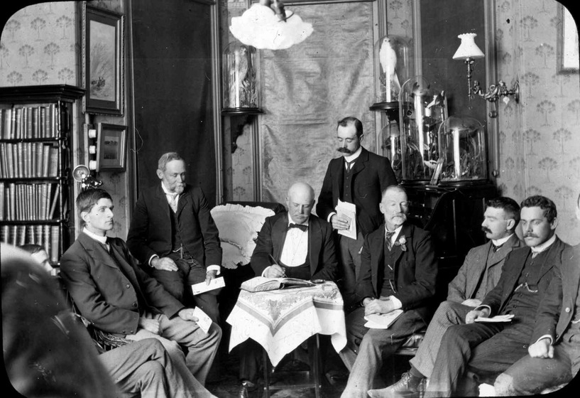 A group of formally-dressed gentlemen meeting around a table within a library, with one man taking notes.