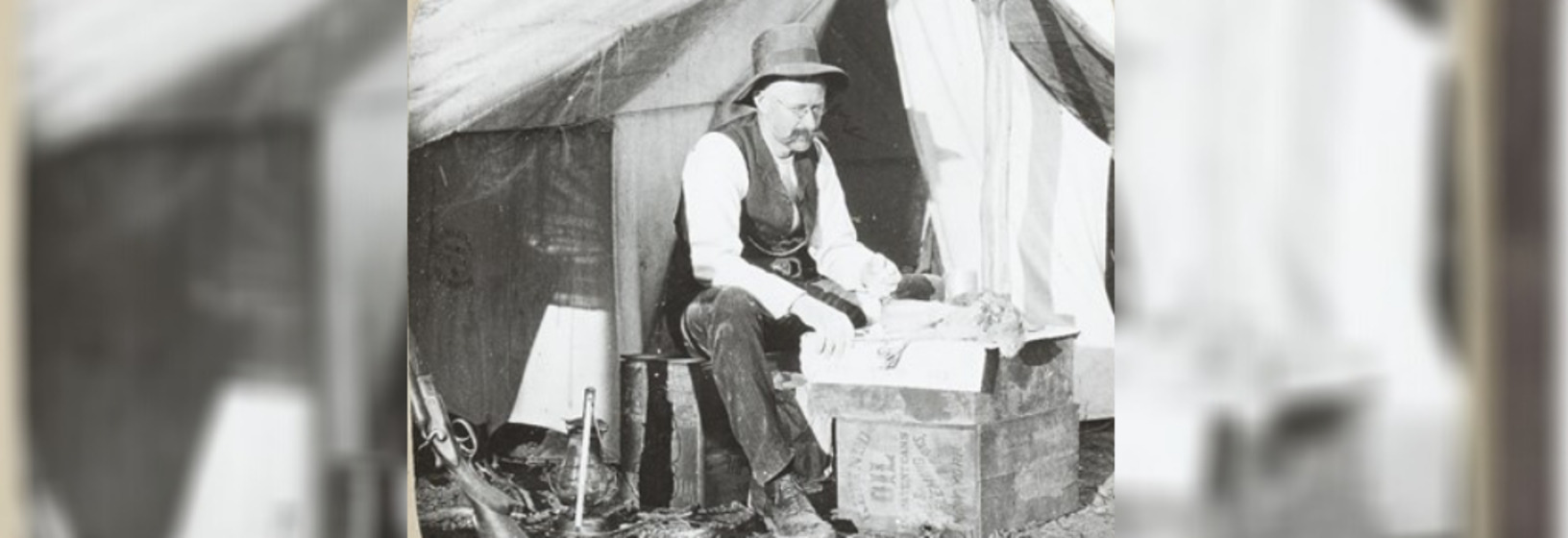 a workman sits on a wooden crate outside a tent, examining bird specimens