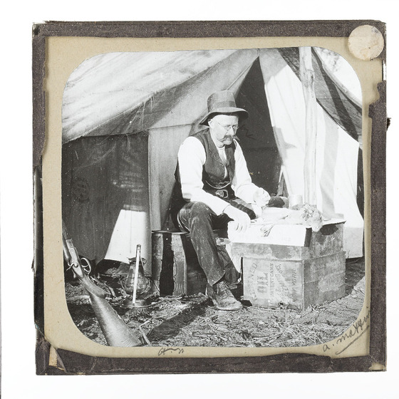 a workman sits on a wooden crate outside a tent, examining bird specimens for field research.