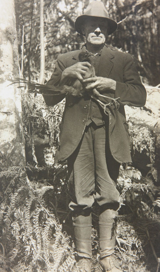 A man dressed all in a suit and hat standing in the forest holding a lyrebird with both hands.