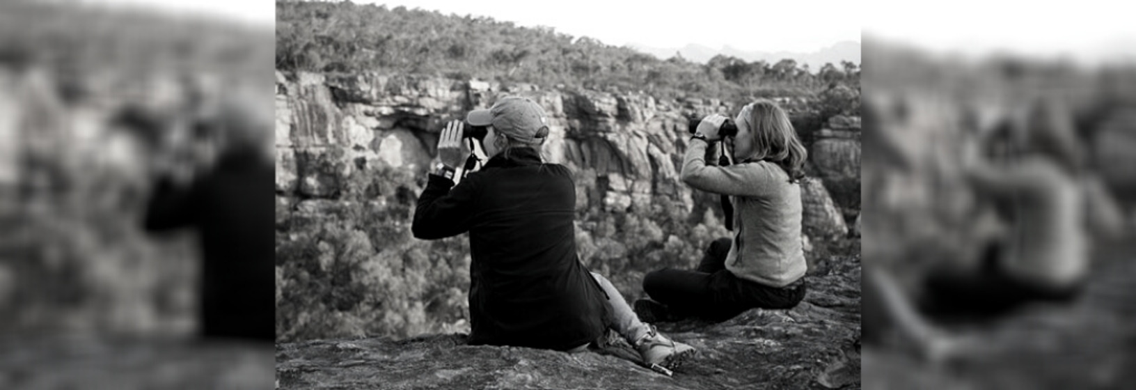 Two women sit high up on a rock and look out into the sprawling landscape through binoculars.