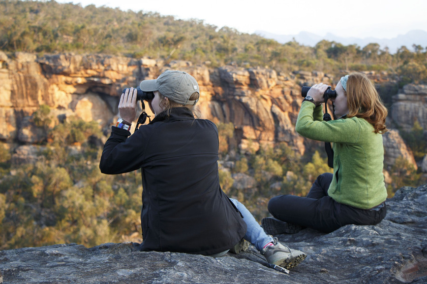 Two women sit high up on a rock and look out into the sprawling landscape through binoculars.