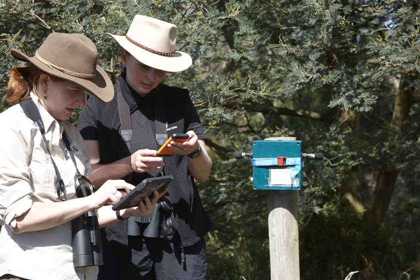 Two women wearing bushman hats holding recording devices stand next to a blue box fitted with two small microphones