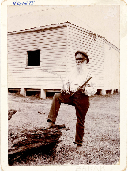man stands with foot raised on log and two Aboriginal weapons in hand