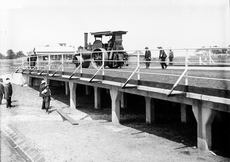 men observe a steam-driven machine crossing a bridge