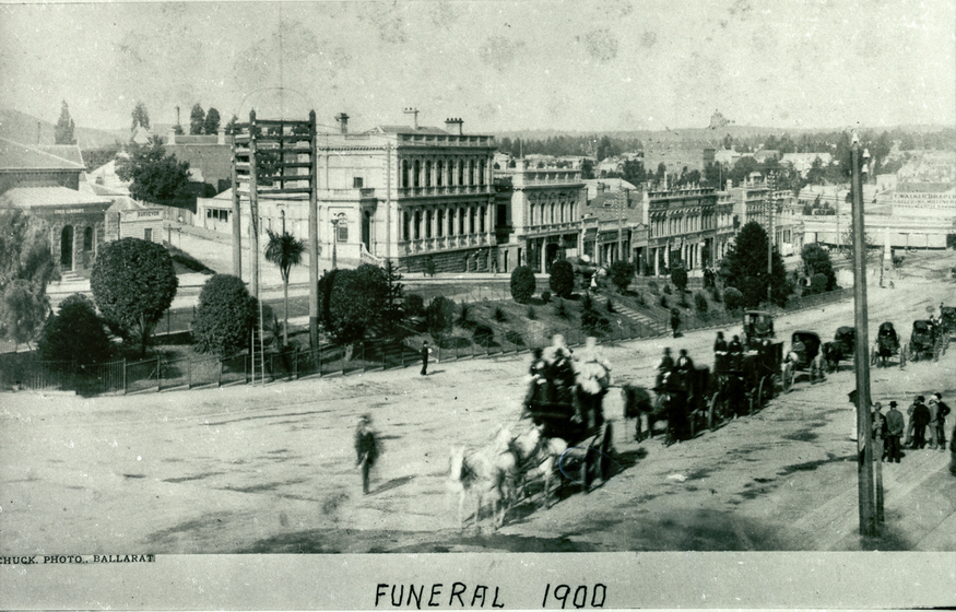 A horse drawn funeral procession and mourners wend their way down the main street