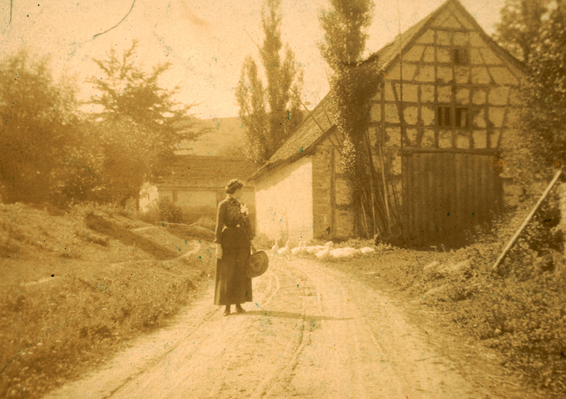 A young woman stands in front of a farmhouse with geese in the background