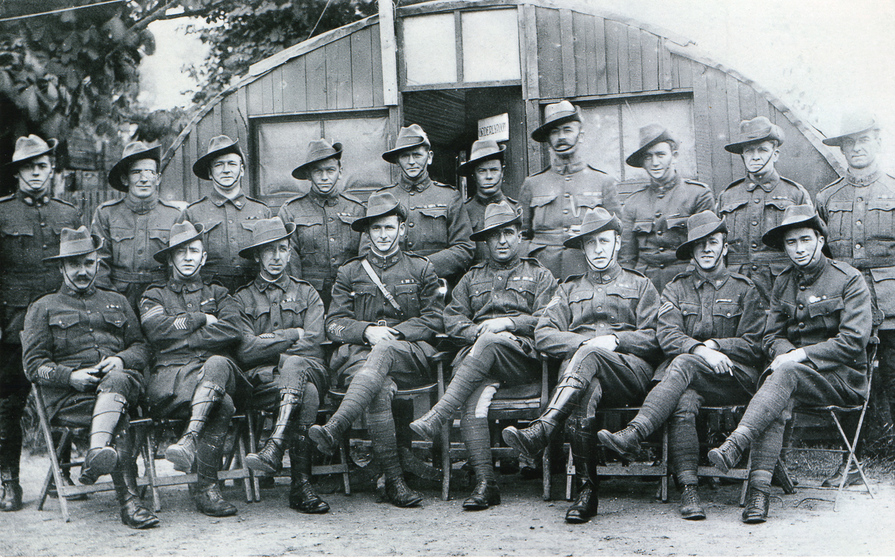 A group of men in uniform in front of a hut, front row seated with legs crossed, back row standing.