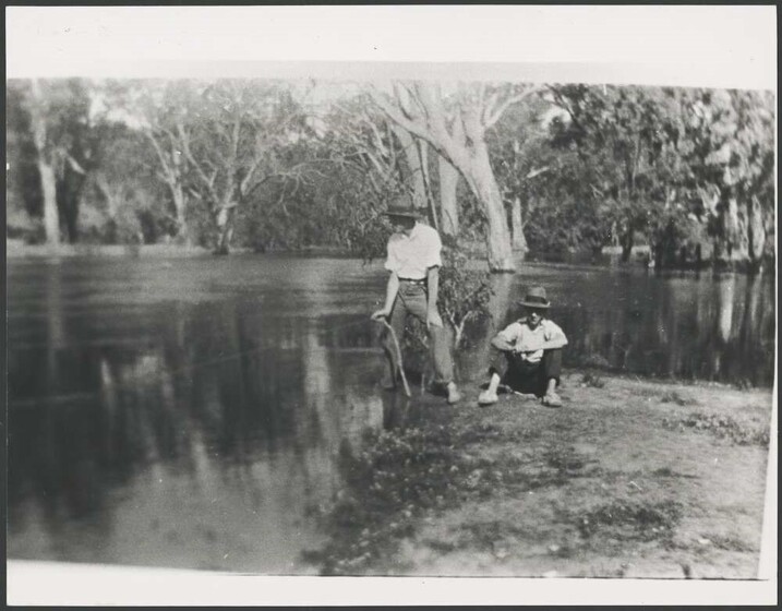 A black and white photograph of two men standing beside a river, holding handmade fishing rods.