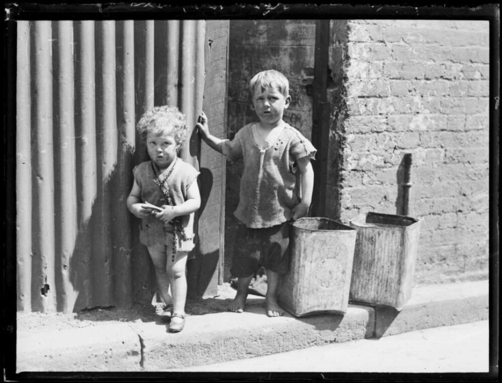 A black and white photograph of two young children in dirty clothing standing on the street.
