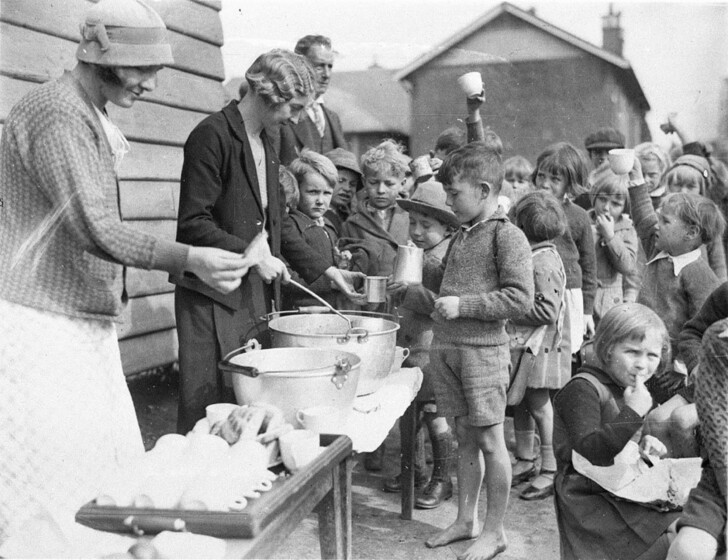 A black and white photograph of children lining up to receive free soup. 
