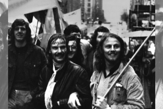 Detail of black and white photograph of demonstrators with banners in a city street, including two men arm in arm, collaged to create a banner image.